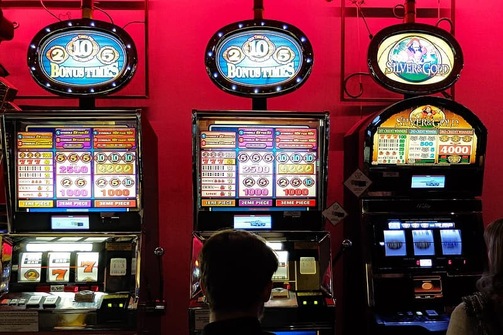 Three slot machines set against a red backdrop with two players in front who have kept their gambling money separate to cash funds