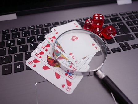 A ten, jack, queen, king, and ace of hearts rest on the keyboard of a silver laptop, with four red dice to the right of the ace of hearts and magnifying glass over the ace